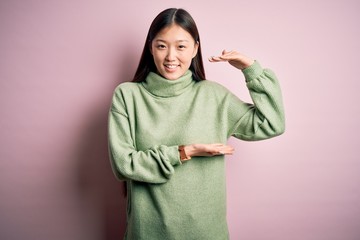 Fototapeta premium Young beautiful asian woman wearing green winter sweater over pink solated background gesturing with hands showing big and large size sign, measure symbol. Smiling looking at the camera. Measuring