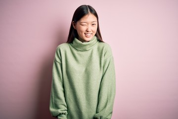 Young beautiful asian woman wearing green winter sweater over pink solated background winking...