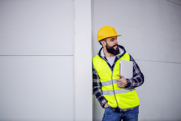 Young smiling attractive caucasian bearded construction worker with helmet on head in vest leaning on the wall, holding laptop in hands and looking at other workers.