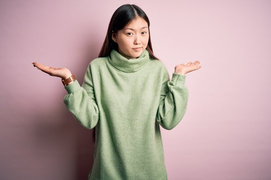 Young beautiful asian woman wearing green winter sweater over pink solated background clueless and confused expression with arms and hands raised. Doubt concept.