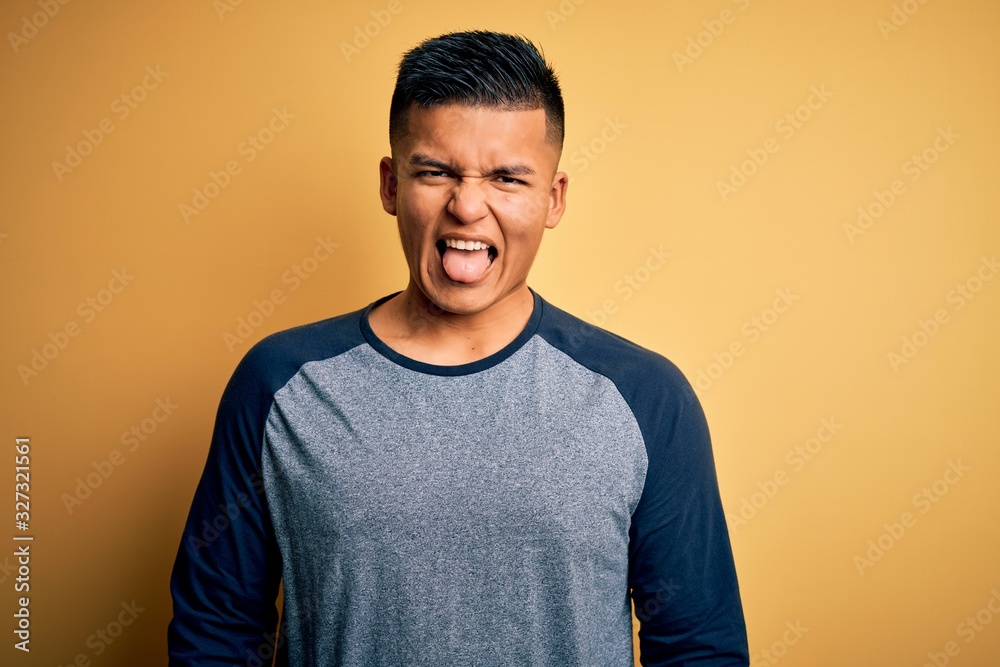 Wall mural young handsome latin man wearing casual t-shirt standing over yellow background sticking tongue out 