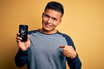 Young handsome latin man holding broken smartphone showing craked screen with surprise face...