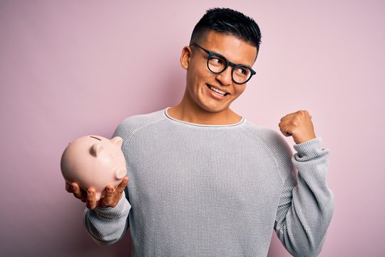 Young handsome latin man holding piggy bank over isolated pink background pointing and showing with thumb up to the side with happy face smiling