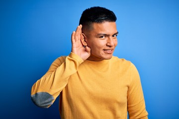 Naklejka premium Young handsome latin man wearing yellow casual sweater over isolated blue background smiling with hand over ear listening an hearing to rumor or gossip. Deafness concept.