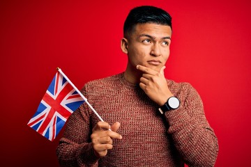 Young handsome latin patriotic man wearing united kingdom flag over red background serious face...