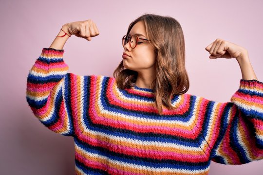 Young beautiful blonde girl wearing glasses and casual sweater over pink isolated background showing arms muscles smiling proud. Fitness concept.