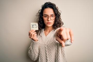 Young beautiful woman with curly hair holding reminder paper with yes word message pointing with...