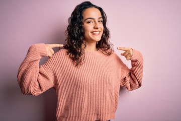 Young beautiful woman with curly hair wearing casual sweater over isolated pink background looking confident with smile on face, pointing oneself with fingers proud and happy.