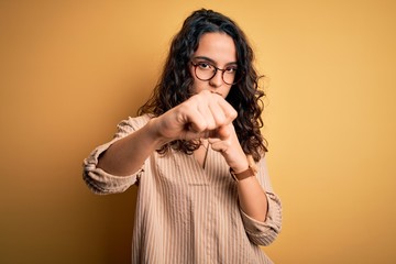 Beautiful woman with curly hair wearing striped shirt and glasses over yellow background Punching fist to fight, aggressive and angry attack, threat and violence