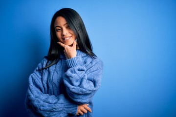 Young beautiful chinese woman wearing casual sweater over isolated blue background looking confident at the camera smiling with crossed arms and hand raised on chin. Thinking positive.