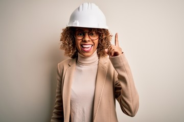 Young african american curly architect woman wearing safety helmet and glasses pointing finger up with successful idea. Exited and happy. Number one.