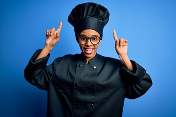 Young african american chef woman wearing cooker uniform and hat over blue background smiling amazed and surprised and pointing up with fingers and raised arms.