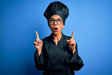 Young african american chef woman wearing cooker uniform and hat over blue background amazed and surprised looking up and pointing with fingers and raised arms.
