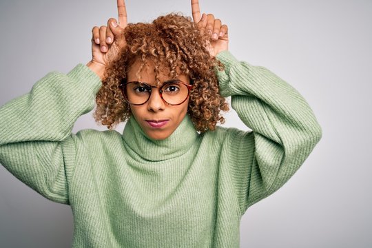Young beautiful african american woman wearing turtleneck sweater and glasses doing funny gesture with finger over head as bull horns