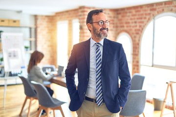 Middle age handsome businessman wearing glasses   standing at the office looking away to side with smile on face, natural expression. Laughing confident.