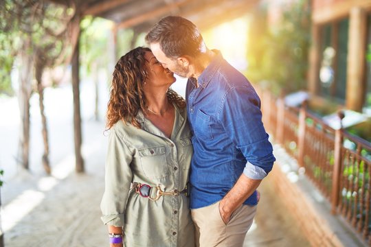 Middle age beautiful couple smiling happy and confident at town park. Standing with smile on face hugging and kissing