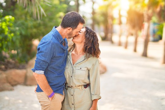 Middle age beautiful couple smiling happy and confident at town park. Standing with smile on face hugging and kissing