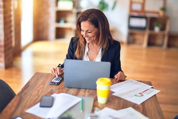 Middle age beautiful businesswoman smiling happy and confident. Sitting on chair working in a desk using calculator at the office