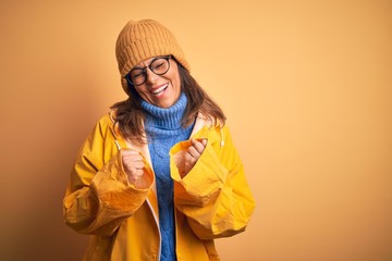 Middle age woman wearing yellow raincoat and winter hat over isolated background excited for success with arms raised and eyes closed celebrating victory smiling. Winner concept.