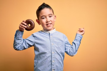 Young little boy kid eating unhealthy chocolate doughnut over isolated yellow background very happy pointing with hand and finger to the side