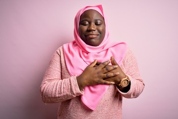 Young african american plus size woman wearing muslim hijab over isolated pink background smiling with hands on chest with closed eyes and grateful gesture on face. Health concept.