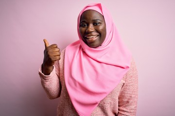 Young african american plus size woman wearing muslim hijab over isolated pink background smiling with happy face looking and pointing to the side with thumb up.