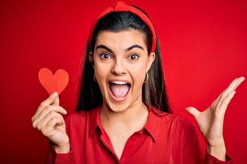 Young beautiful brunette romantic woman holding red heart paper for valentine day very happy and excited, winner expression celebrating victory screaming with big smile and raised hands
