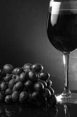 A glass of red wine next to grapes on a dark background. Vertical photo. Close-up. Black and white photography