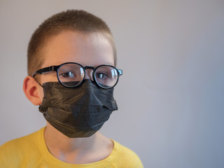 Fototapeta na wymiar cute blonde boy in black medical mask and glasses is quarantined at home. child coughs heavily and wears mask. concept of fight against the coronavirus epidemic and proper prevention of infections