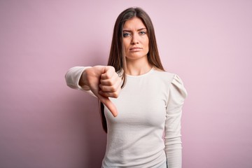 Fototapeta premium Young beautiful woman with blue eyes wearing casual white t-shirt over pink background looking unhappy and angry showing rejection and negative with thumbs down gesture. Bad expression.
