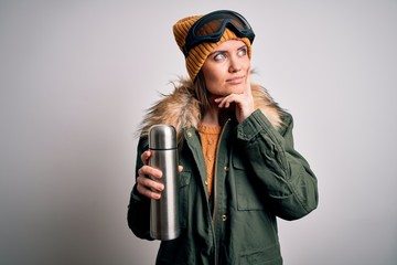 Young beautiful skier woman with blue eyes wearing ski goggles drinking thermo with coffee serious face thinking about question, very confused idea