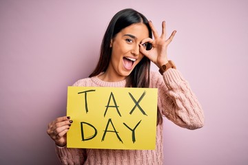 Young beautiful brunette woman holding paper with tax day message over pink background with happy face smiling doing ok sign with hand on eye looking through fingers