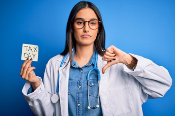 Young doctor woman wearing stethoscope holding paper with tax day message with angry face, negative sign showing dislike with thumbs down, rejection concept