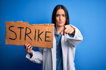 Young doctor woman wearing stethoscope holding cardboard banner protesting in strike pointing with finger to the camera and to you, hand sign, positive and confident gesture from the front