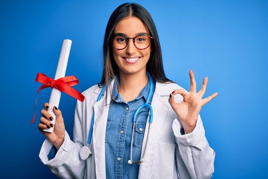 Young beautiful brunette doctor woman wearing glasses and coat holding diploma degree doing ok sign with fingers, excellent symbol