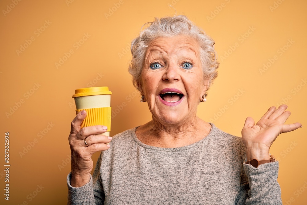 Wall mural senior beautiful woman drinking cup of coffee standing over isolated yellow background very happy an