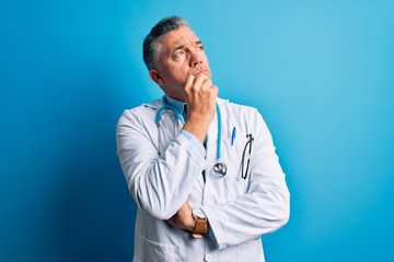 Middle age handsome grey-haired doctor man wearing coat and blue stethoscope with hand on chin...