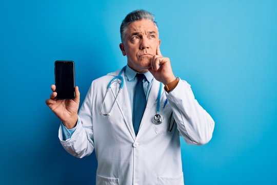 Middle age handsome grey-haired doctor man holding smartphone showing screen serious face thinking about question, very confused idea