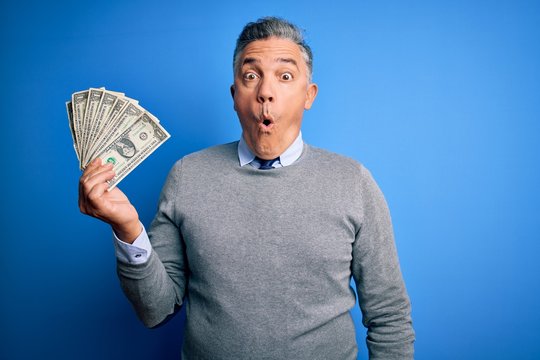 Middle age handsome grey-haired man holding bunch of dollars over blue background scared in shock with a surprise face, afraid and excited with fear expression