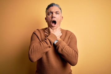 Middle age handsome grey-haired man wearing casual sweater over yellow background shouting and suffocate because painful strangle. Health problem. Asphyxiate and suicide concept.