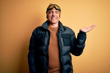 Middle age handsome grey-haired skier man on vacation wearing ski goggles smiling cheerful presenting and pointing with palm of hand looking at the camera.