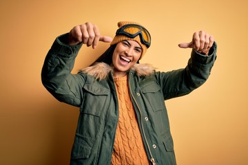 Young beautiful hispanic woman wearing ski glasses and coat for winter weather approving doing positive gesture with hand, thumbs up smiling and happy for success. Winner gesture.