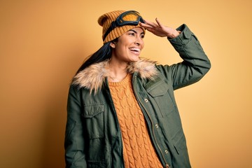 Young beautiful hispanic woman wearing ski glasses and coat for winter weather very happy and smiling looking far away with hand over head. Searching concept.