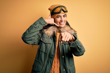 Young beautiful hispanic woman wearing ski glasses and coat for winter weather smiling doing talking on the telephone gesture and pointing to you. Call me.