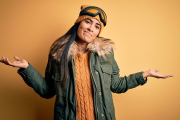 Young beautiful hispanic woman wearing ski glasses and coat for winter weather clueless and confused expression with arms and hands raised. Doubt concept.