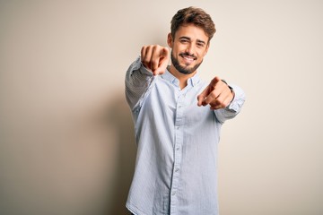Young handsome man with beard wearing striped shirt standing over white background pointing to you and the camera with fingers, smiling positive and cheerful