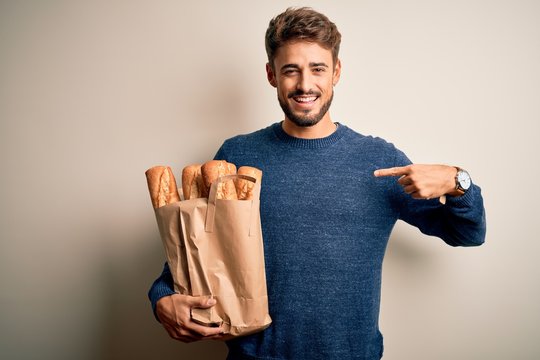 Young man holding paper bag with bread standing over isolated red bakground with surprise face pointing finger to himself