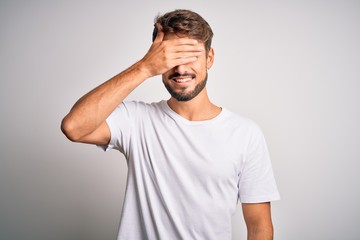 Fototapeta na wymiar Young handsome man with beard wearing casual t-shirt standing over white background smiling and laughing with hand on face covering eyes for surprise. Blind concept.