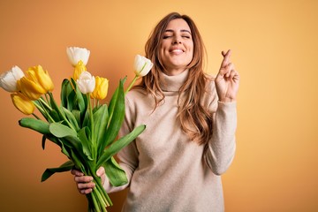 Young beautiful brunette woman holding bouquet of yellow tulips over isolated background gesturing finger crossed smiling with hope and eyes closed. Luck and superstitious concept.