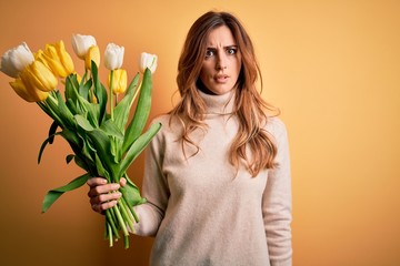 Young beautiful brunette woman holding bouquet of yellow tulips over isolated background skeptic...